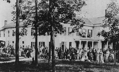 Historic photograph of the new Western Health Reform Institute building in the mid-1870s, with Mrs. White in reform dress and Elder White standing to the right of trees